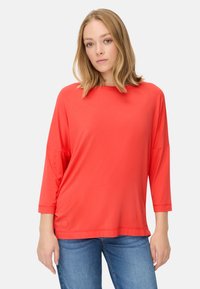 Rød, letvægts, langærmet top med rundt halsudskæring, løs pasform og sidelæg, parret med blå jeans.