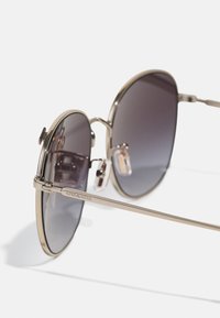 Óculos de sol em forma redonda com armação dourada metálica e lentes de gradiente cinza-escuras, apresentando uma ponte dupla e a gravação "COACH" nas hastes.
