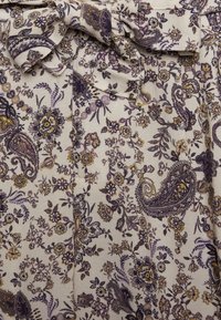 Tecido com padrão floral em creme, apresentando desenhos de paisley e flores nas cores roxo, amarelo e preto. Detalhe de laço na parte superior.