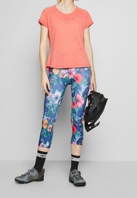 Koraalkleurig T-shirt met korte mouwen en knoopdetails, gecombineerd met bloemenpatroon leggings in blauwe, roze en groene tinten. Inclusief grijze sportschoenen.