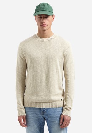 Man draagt een beige gebreide trui, lichtblauwe spijkerbroek en een groene baseballpet, staand tegen een effen witte achtergrond.