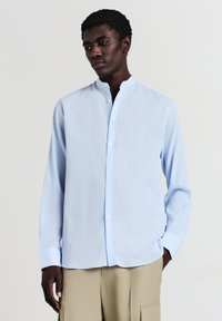 Camicia azzurra, a maniche lunghe, con colletto alla coreana e una texture sottile. Presenta una pattina con bottoni e maniche con polsini. Tessuto leggero.