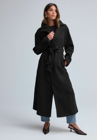 Trench coat dintr-un amestec de lână neagră, cu un guler larg, talie cu brâu, mâneci lungi și buzunare laterale, asortat cu blugi scurți și pantofi cu toc negri.