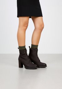 Botas de tobillo marrones con cordones, con un tacón grueso y una suela con relieve, que presentan calcetines verdes asomando por la parte superior. Material suave con cordones decorativos.