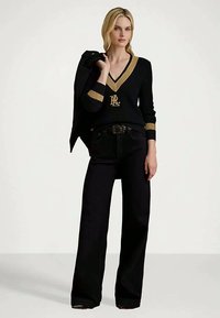 Ξανθιά γυναίκα που φοράει μαύρο παντελόνι φαρδύ και μαύρο πουλόβερ με χρυσές λεπτομέρειες, κρατώντας μια μαύρη ζακέτα πάνω στον ώμο της.