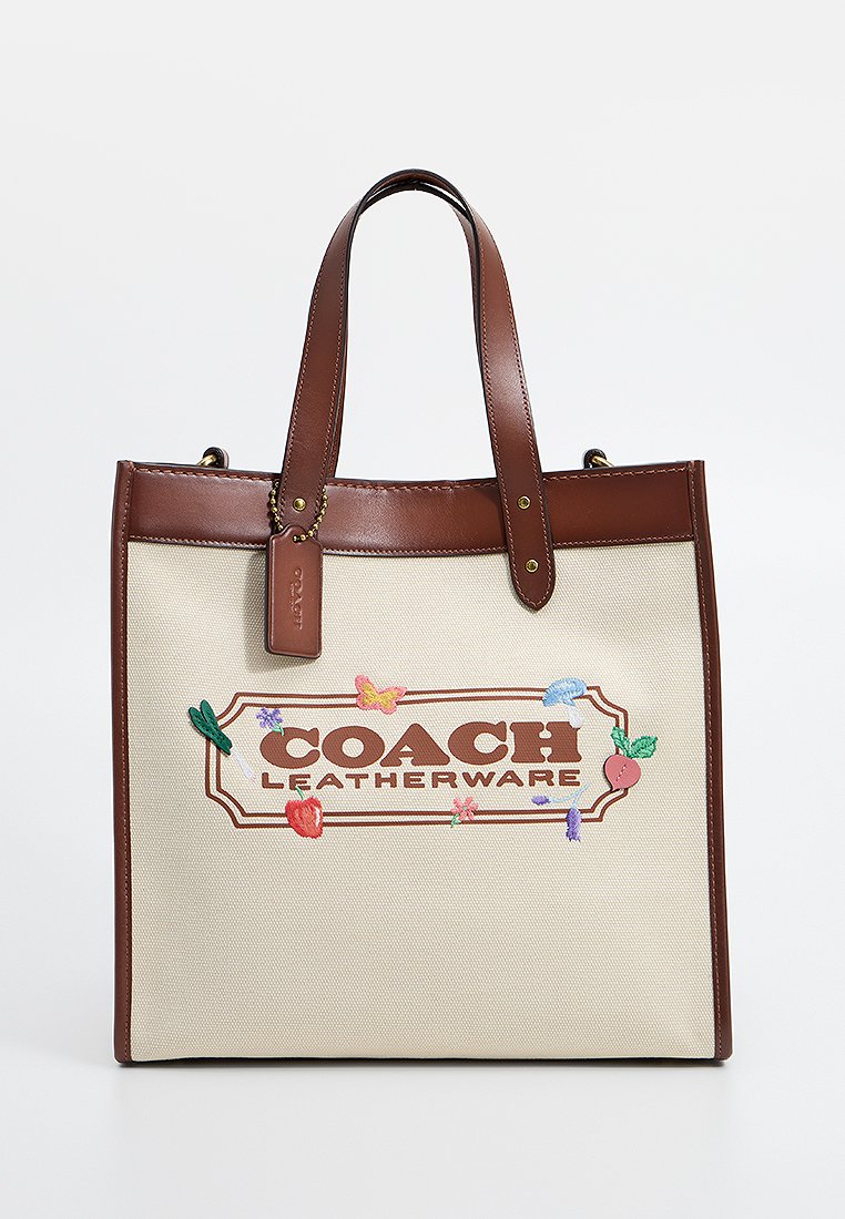 Bolsa de lona beige con asas y ribetes de cuero marrón. Presenta diseños bordados de flores y mariposas, y el logo "COACH LEATHERWARE".
