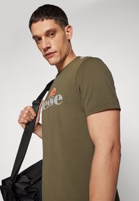 Olive green T-shirt with "ellesse" logo in grey, short sleeves, held by a person carrying a black shoulder bag, plain background.