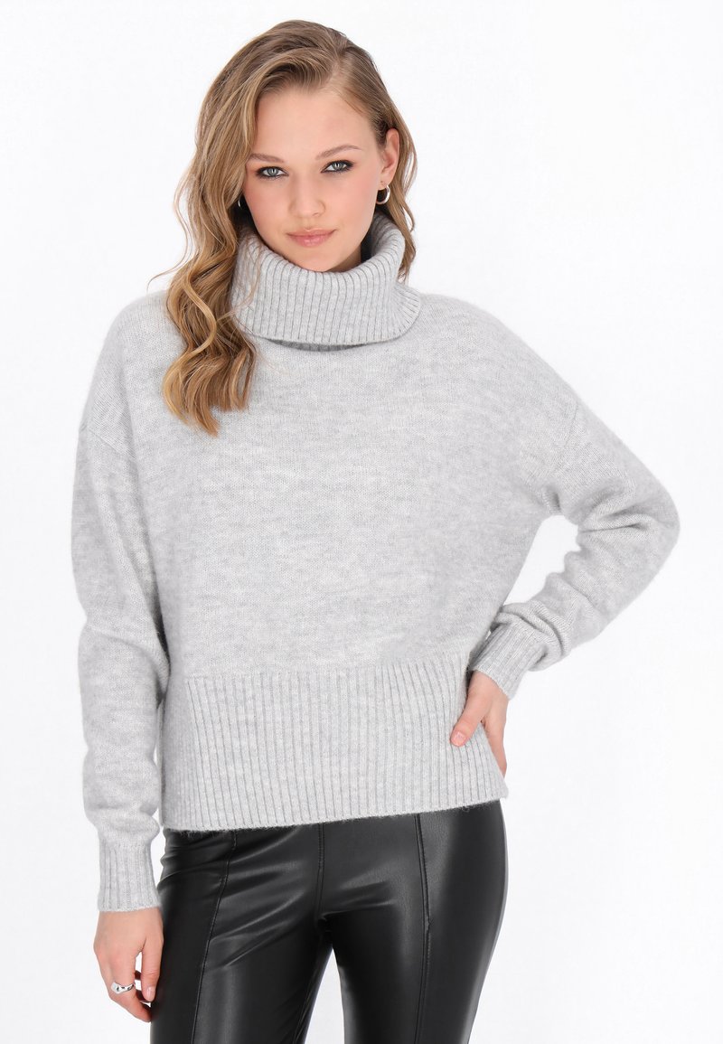 Helles graues Strickpullover mit hohem, geripptem Rollkragen und geripptem Saum, mit weit geschnittenen Ärmeln und weichem Material. Kombiniert mit schwarzen Hosen.