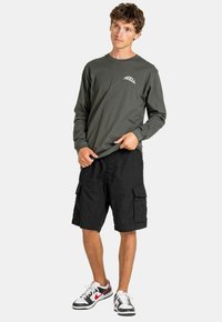 Camisa de manga larga verde oliva con el logotipo "REELL", pantalones cortos cargo negros y zapatillas en blanco y negro. Tejido texturizado y ajuste relajado.