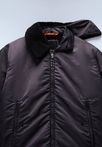 Giacca nera imbottita con colletto in pile strutturato, zip centrale, tasche con bottone a pressione e risvolto del colletto piegato.