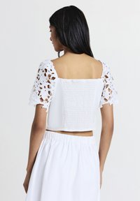 Femme portant un haut blanc court avec des manches en dentelle florale et un corsage froncé, associé à une jupe blanche avec taille élastique, tournant le dos.