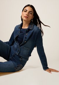 Veste en jean avec un col orné, présentant un design ajusté, des boutons sur le devant, et associée à un jean taille haute avec broderies florales.