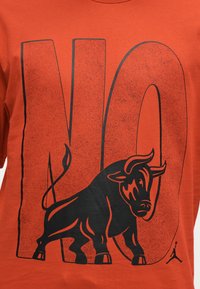 Μεγάλο μαύρο γραφικό ταύρου που επικαλύπτει έντονο, φθαρμένο κείμενο "NO" σε πορτοκαλί μπλουζάκι, με μικρό λογότυπο Jumpman στη κάτω δεξιά γωνία.