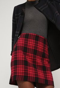 Rode en zwarte geruite mini-rok met een rechte snit, voorzien van ritsen aan de zijkanten, gecombineerd met een grijze coltrui en een patroonjas.