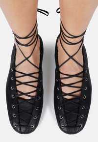 Ballerines noires à lacets avec une finition en cuir lisse, présentant des lacets entrecroisés à travers des œillets en métal et des liens autour de la cheville, offrant un design minimaliste.