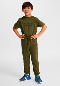 Conjunto de chándal verde oliva con camiseta de manga corta y pantalones con cintura elástica. Diseño ajustado con detalle de logo "hummel". Zapatillas azules.