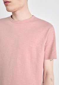 Pale rózsaszín, rövid ujjú póló puha, texturált anyagból, diszkrét domború logóval a mellrészen és kerek nyakkivágással.