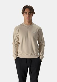 Felpa beige con scollatura rotonda, maniche lunghe e dettaglio logo ricamato. Tessuto liscio, silhouette aderente, abbinata a pantaloni neri.
