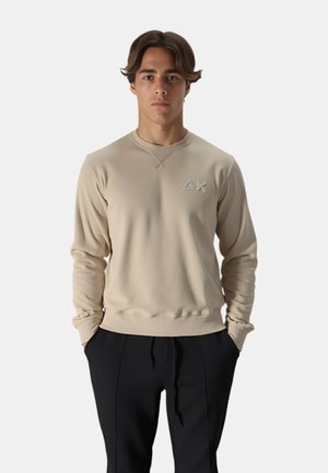 Felpa beige con scollatura rotonda, maniche lunghe e dettaglio logo ricamato. Tessuto liscio, silhouette aderente, abbinata a pantaloni neri.