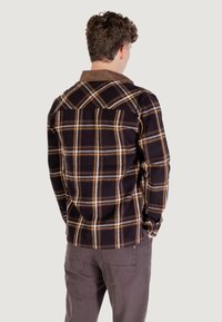Koszula flannel w kratę w ciemnym brązie z żółtymi i białymi akcentami. Posiada kontrastowy kołnierz oraz detale na yoku pleców.
