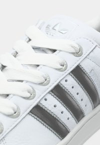 Gros plan sur une sneaker Adidas blanche avec des lacets blancs, des rayures noires, des œillets en métal et un logo en relief de la trêfle Adidas sur la languette.