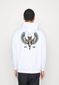 Uomo che indossa una felpa bianca con cappuccio con un disegno colorato di un gatto nero alato e il testo "est. 1990" sul retro, in piedi davanti a uno sfondo bianco semplice.