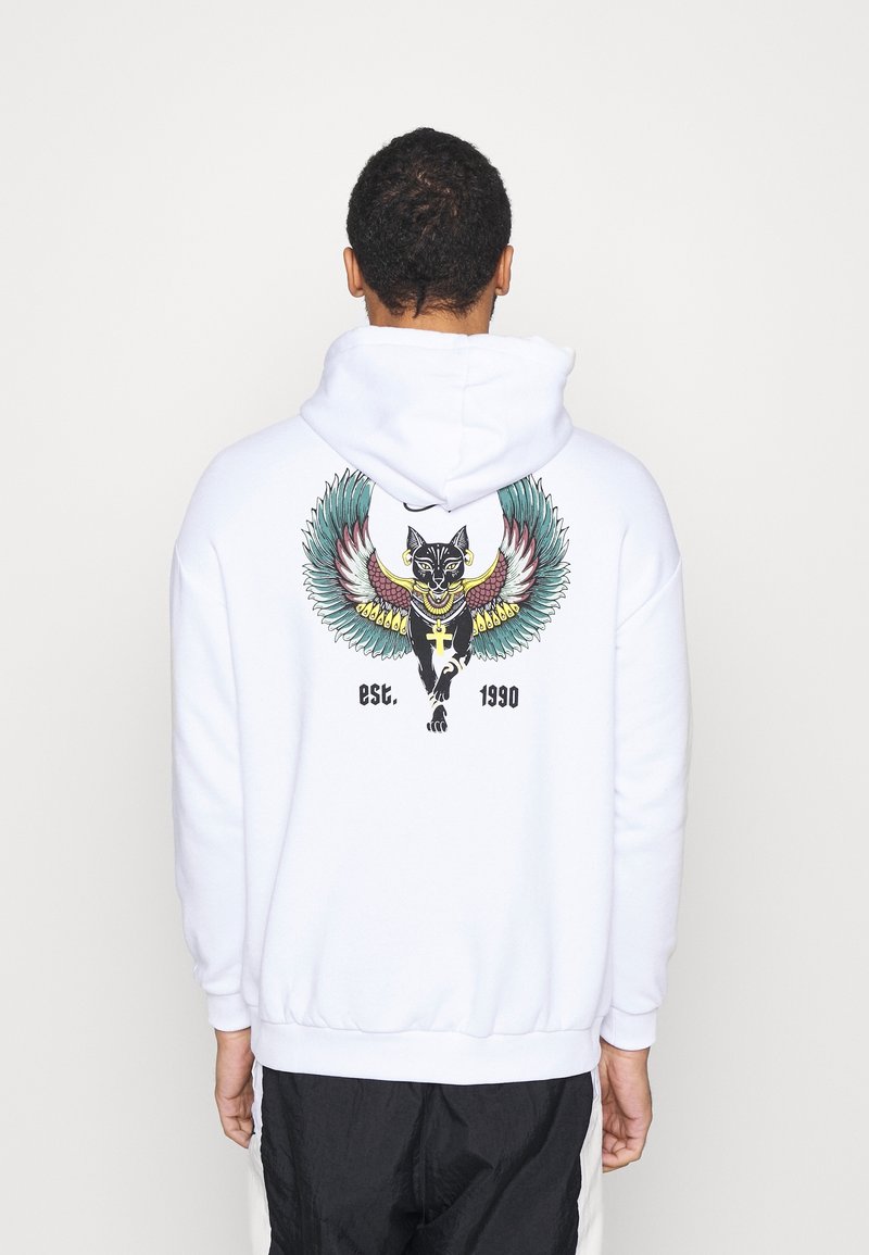 Uomo che indossa una felpa bianca con cappuccio con un disegno colorato di un gatto nero alato e il testo "est. 1990" sul retro, in piedi davanti a uno sfondo bianco semplice.