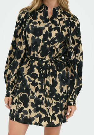 Femme portant une robe beige avec un grand imprimé floral noir, manches longues bouffantes, boutonnée sur le devant, et une ceinture en tissu nouée à la taille.