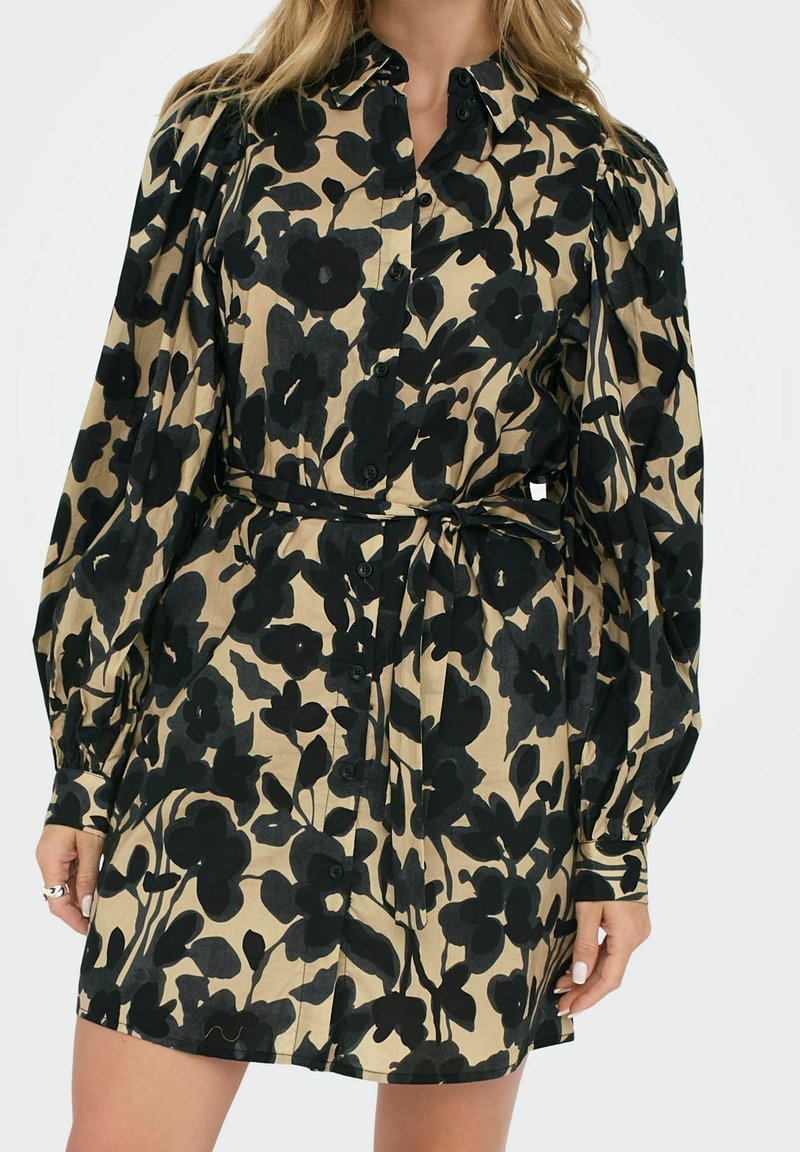 Femme portant une robe beige avec un grand imprimé floral noir, manches longues bouffantes, boutonnée sur le devant, et une ceinture en tissu nouée à la taille.