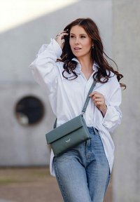 Zelená kožená crossbody taška s překlápěcí klopou, čistými liniemi a jemným brandingem. Nosená s bílou košile a modrými džíny.