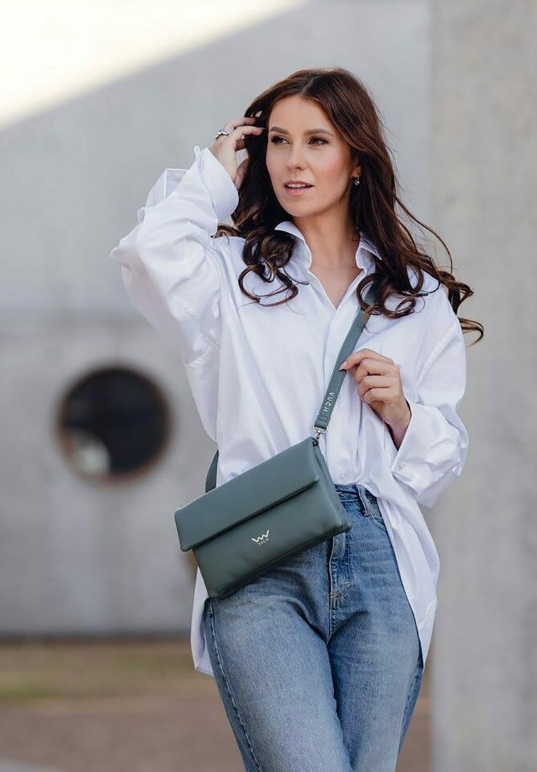 Zelená kožená crossbody taška s překlápěcí klopou, čistými liniemi a jemným brandingem. Nosená s bílou košile a modrými džíny.