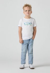 Fehér, rövid ujjú póló színes Levi's logó grafikával, világoskék farmernadrággal és szürke részletekkel ellátott fehér tornacipővel párosítva.