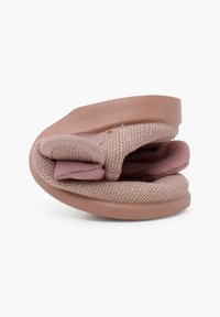 Zapatilla de color rosa suave, sin cordones, con una parte superior de tejido texturizado y un acento acolchado. La suela es redondeada y tiene un acabado suave para mayor comodidad.