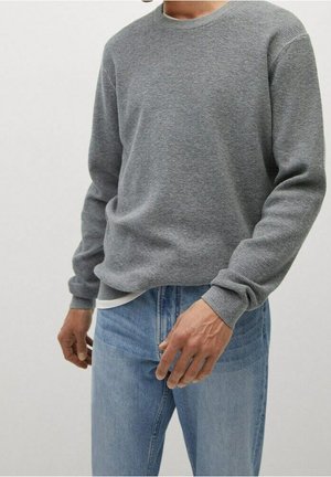 Homme portant un pull gris texturé à manches longues sur une chemise blanche, associé à un jean bleu clair sur un fond uni.