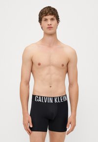 Boxeri scurți negri din bumbac, cu un tiv lat și contrastant cu logo-ul Calvin Klein. Textură netedă, design ajustat și lungime până la mijlocul coapsei.