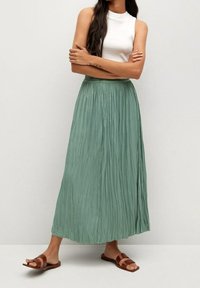 Vrouw met lang donker haar, gekleed in een mouwloze witte top, een mintgroene geplooide maxi-skirt en bruine platte sandalen, staand met de armen over elkaar.