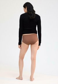 Bruine high-waisted onderbroek gecombineerd met een zwarte longsleeve top. Glad stof, aansluitend ontwerp en geen zichtbare naden. Model kijkt weg.