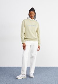 Lys grønn hettegenser med rund hals og brodert "Carhartt"-logo i blått, parret med hvite jeans og grå sneakers.