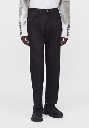 Person iført sorte jeans med lige ben, sorte sneakers og en hvid langærmet skjorte med grå ærmer og tekst på armene.