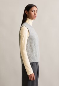 Gilet sans manches gris en matière douce, superposé sur un col roulé beige clair, associé à un pantalon à carreaux dans des tons sombres.