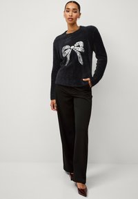 Fuzzy zwarte trui met lange mouwen, voorzien van een grote zilveren strik graphic. Gecombineerd met zwarte wijde broek en puntige bordeauxrode hakken.