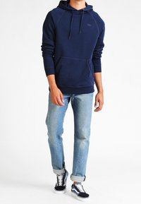 Námořnicky modrá mikina s kapucí, přední kapsou na tlapku, stahovací šňůrkou na kapuci a kontrastním šitím, kombinovaná se světle modrými džínami a černými teniskami.
