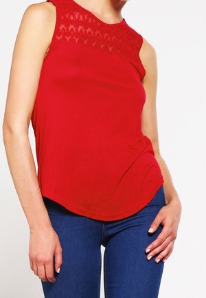 Vrouw in een mouwloze rode top met kantdetails bij de halslijn en blauwe strakke jeans, staand met één hand op de heup.