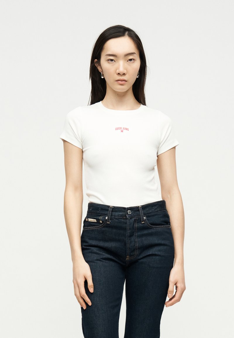 Νεαρή γυναίκα με λευκό εφαρμοστό t-shirt με κόκκινο κείμενο "GUESS JEANS" και σκούρο μπλε ψηλόμεσο τζιν, στέκεται μπροστά σε απλό φόντο.