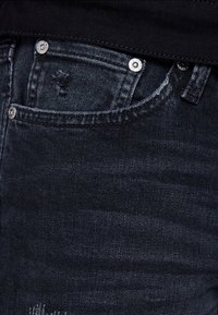Närbild av en mörkblå denimjeans framficka med metallnitar och lätt slitna detaljer nära fickans kant.