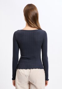 Granatowa bluzka z długim rękawem o ribbowanej strukturze, okrągłym dekolcie, dopasowanym kroju i falbaniastym wykończeniu, zestawiona z jasnymi spodniami w paski.