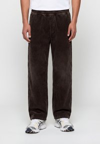 Άνδρας που φοράει παντελόνι από καφέ σκούρο κορδόνι με φαρδύ πόδι και λευκά αθλητικά παπούτσια, στέκεται μπροστά σε ένα απλό φωτεινό φόντο.