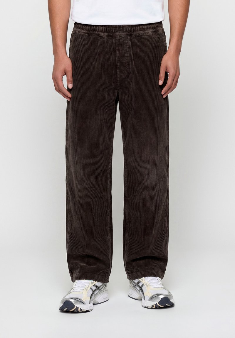 Άνδρας που φοράει παντελόνι από καφέ σκούρο κορδόνι με φαρδύ πόδι και λευκά αθλητικά παπούτσια, στέκεται μπροστά σε ένα απλό φωτεινό φόντο.