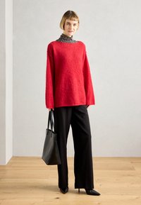 Rød, loddent genser med brede ermer, brukt over en høyhalset topp med svart og hvit stripe. Kombinert med svarte vide bukser og en svart tote bag.