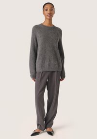 Grå strikket sweater med lange ærmer, parret med grå stribede vidde bukser og sorte spidse sko, sat mod en neutral baggrund.